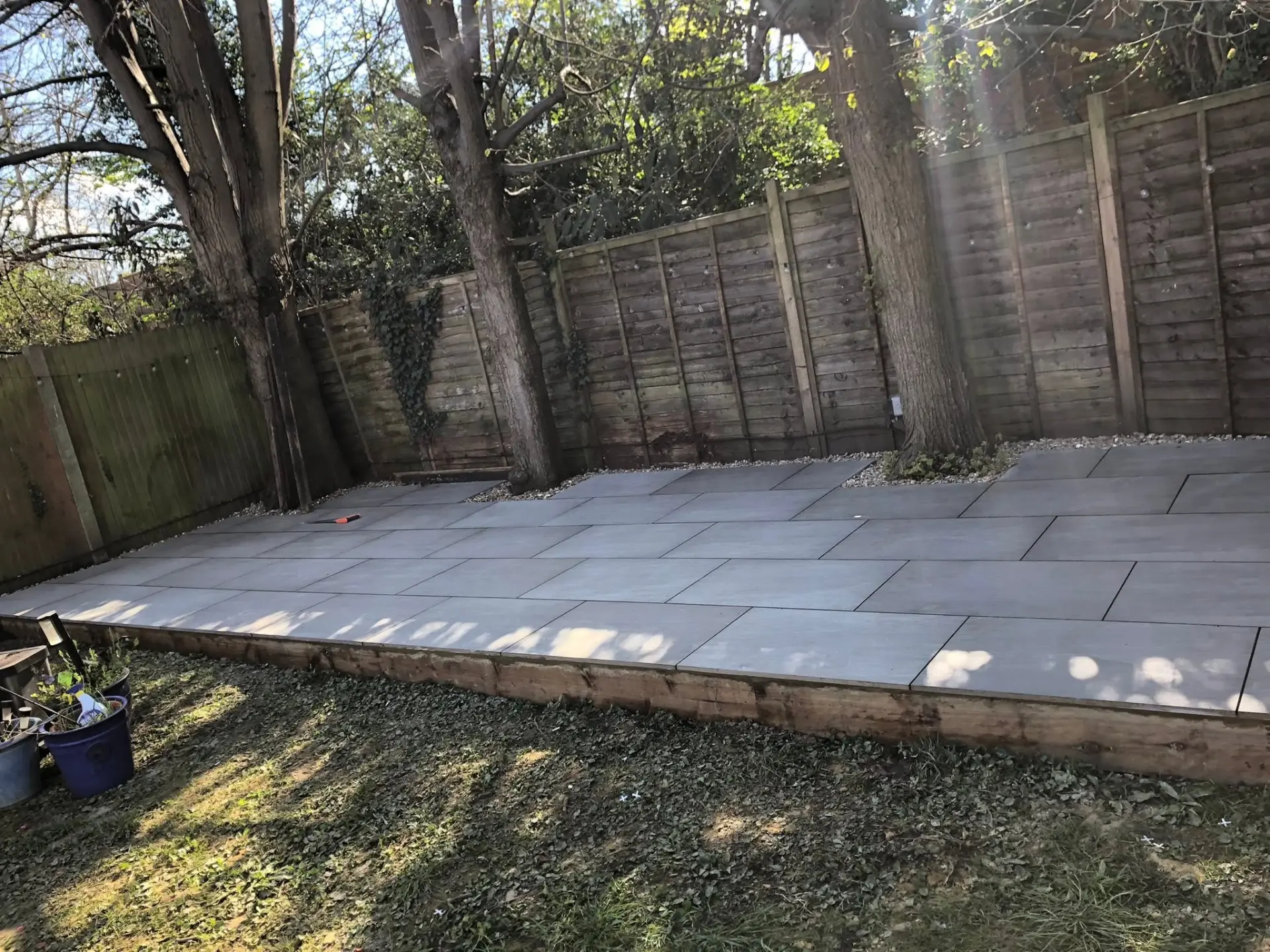 Large grey porcelain patio with timber sleeper edging in a tree-lined rear garden